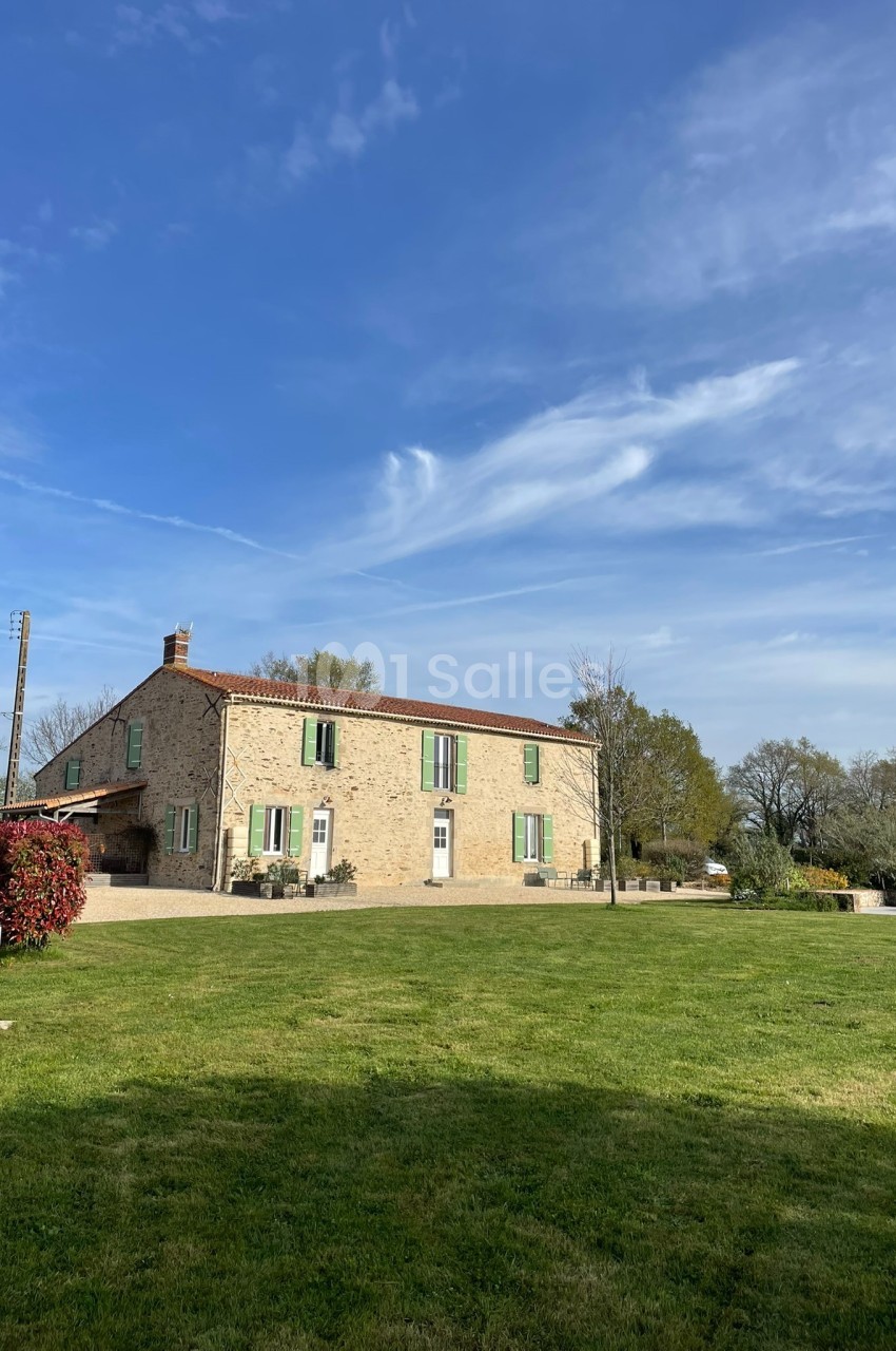 Maison en pierre avec volets verts, entourée d'un grand jardin et d'arbres, sous un ciel bleu dégagé.