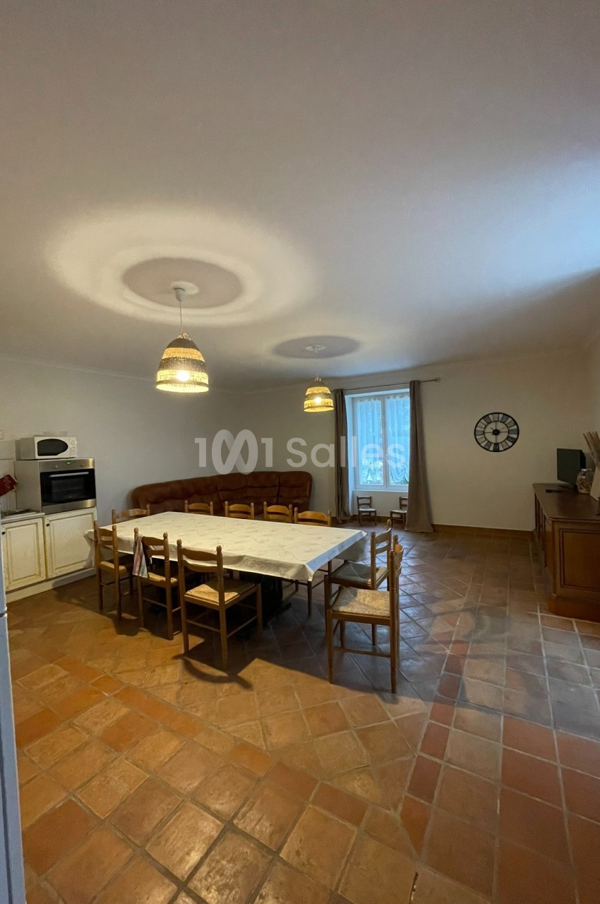 Salle à manger avec une grande table entourée de chaises, carrelage au sol, cuisine équipée et salon en arrière-plan.