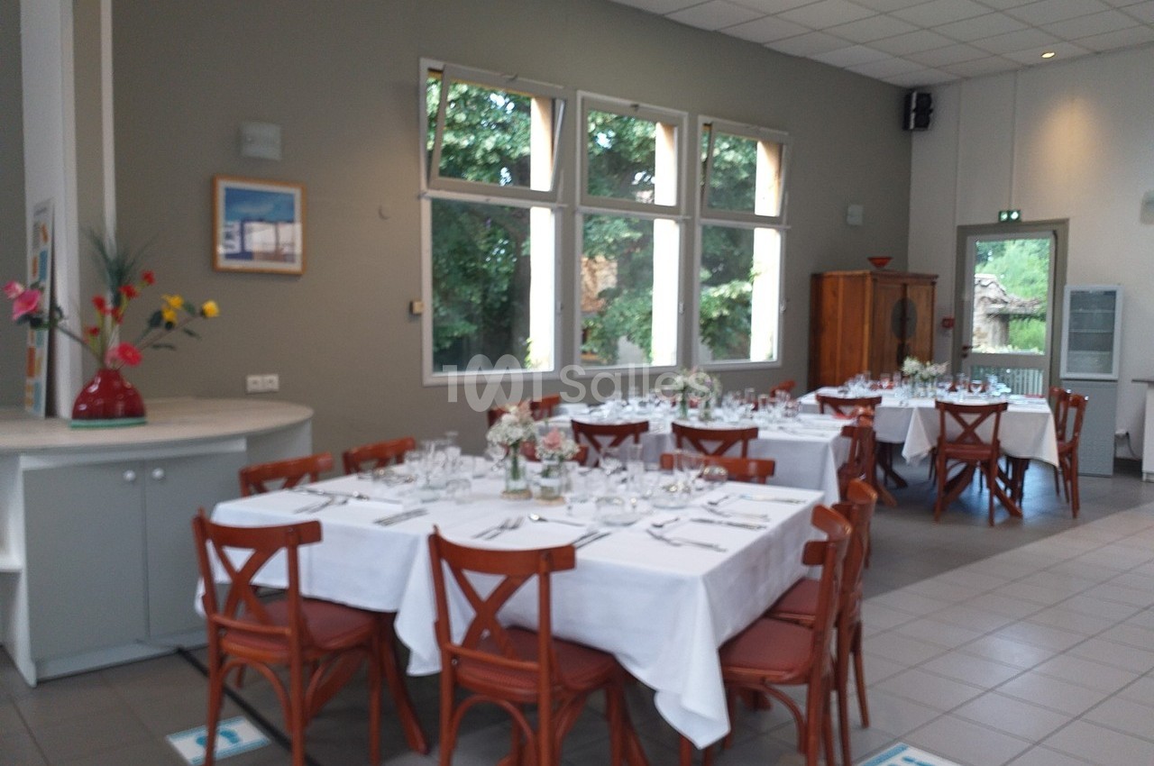 Salle lumineuse avec tables dressées, nappes blanches, chaises en bois et grandes fenêtres donnant sur un jardin.