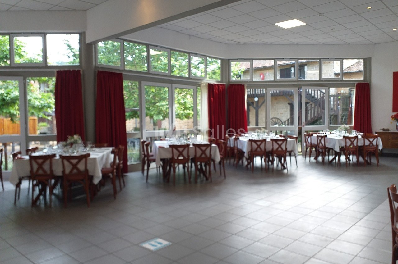 Salle lumineuse avec de grandes baies vitrées, tables rondes dressées avec nappes blanches et rideaux rouges.