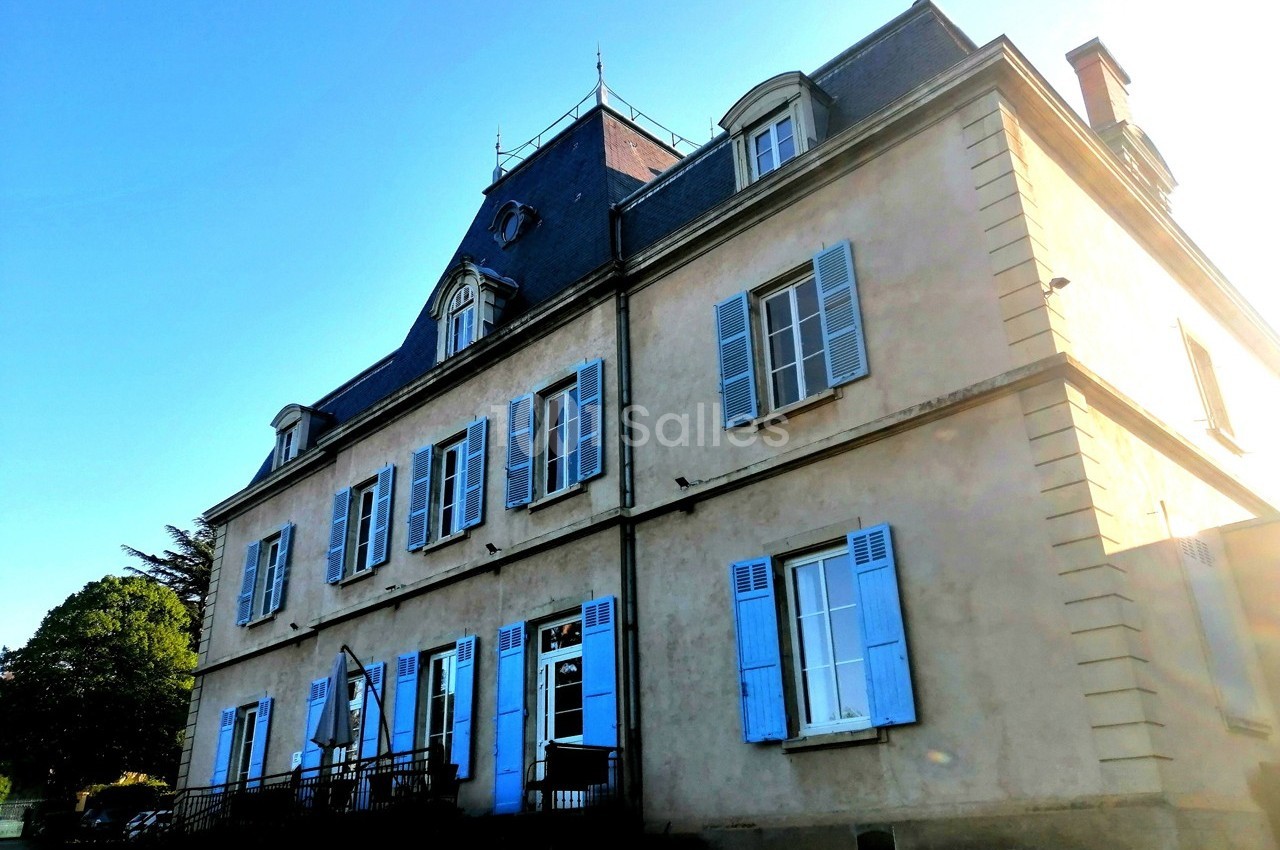 Façade d'une grande maison ancienne avec des volets bleus, un toit en ardoise et un ciel dégagé en arrière-plan.