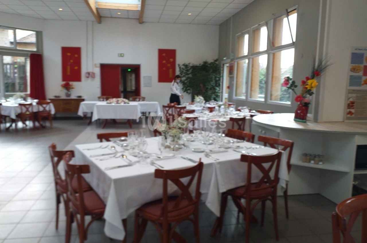 Salle de restaurant lumineuse avec tables dressées de nappes blanches, chaises en bois et décorations florales.