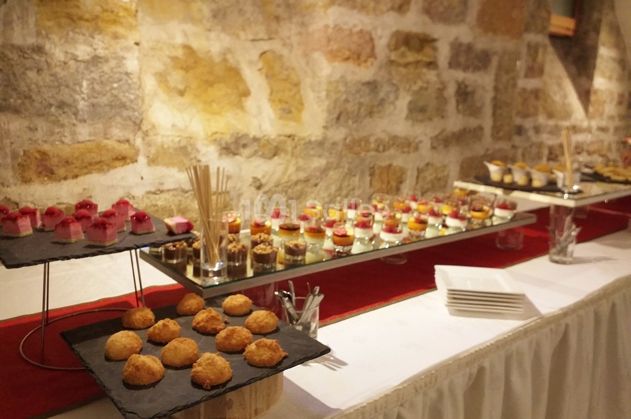 Buffet de desserts variés disposés sur des plateaux devant un mur en pierre, avec nappes blanches et décoration sobre.