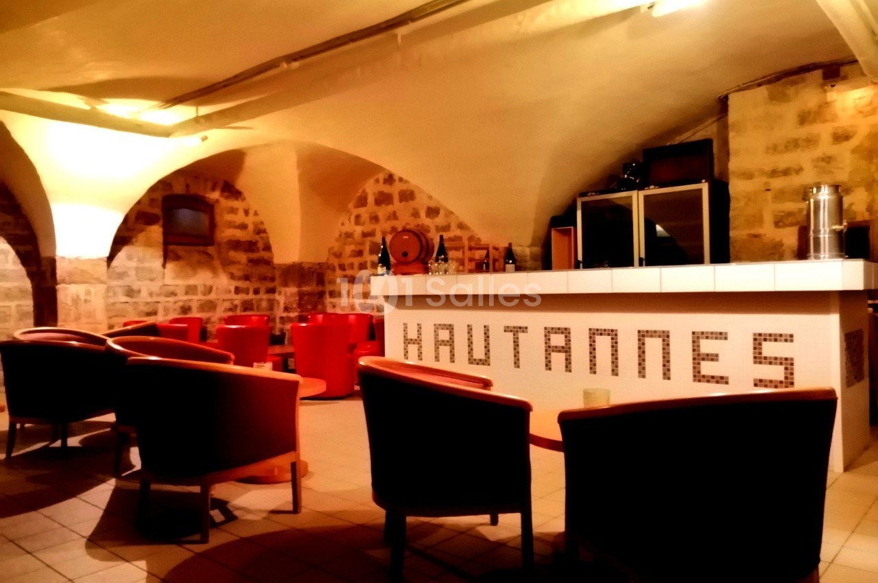 Salle voûtée avec des murs en pierre, un bar blanc avec l'inscription ’Hautannes’ et des fauteuils rouges.