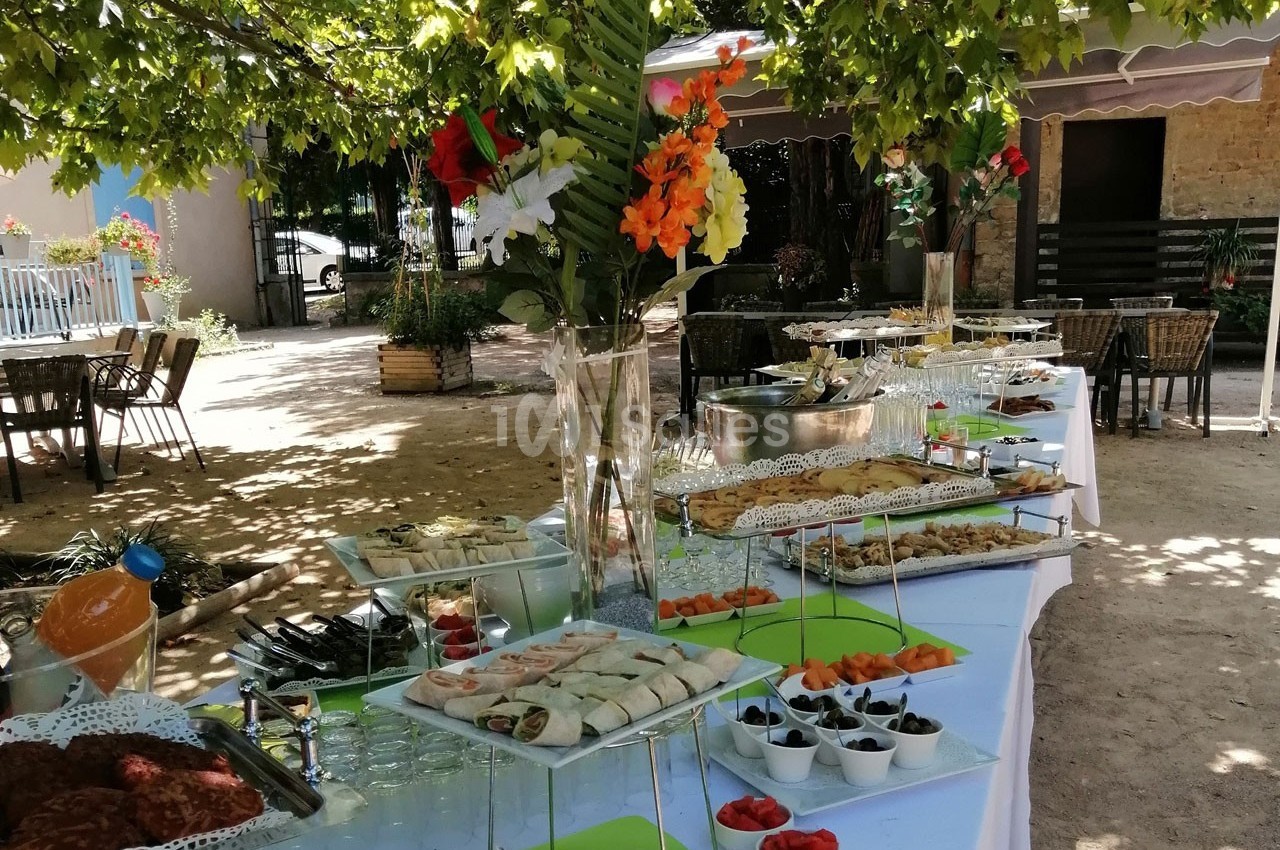 Buffet en extérieur sous des arbres, avec des plats variés, boissons et décorations florales sur des tables dressées.