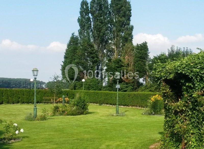Jardin verdoyant avec pelouse, parterres fleuris, lampadaires verts et haies, entouré d'arbres sous un ciel dégagé.