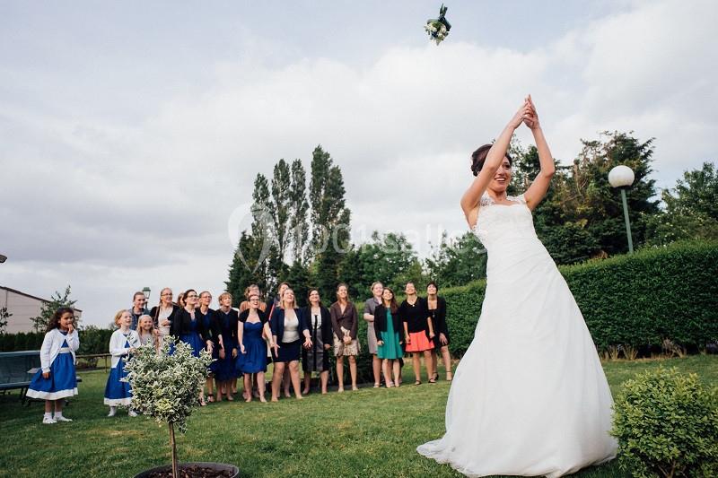 Une mariée lance son bouquet de fleurs vers un groupe de femmes rassemblées dans un jardin.