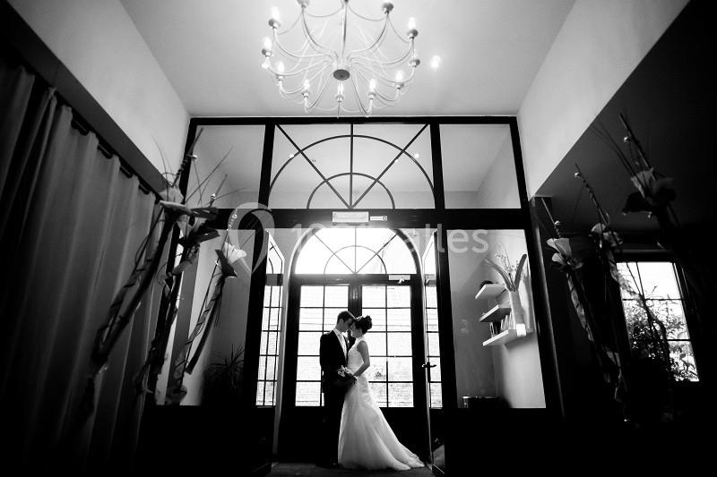 Un couple de mariés s'embrasse sous un lustre dans une entrée lumineuse avec de grandes fenêtres.
