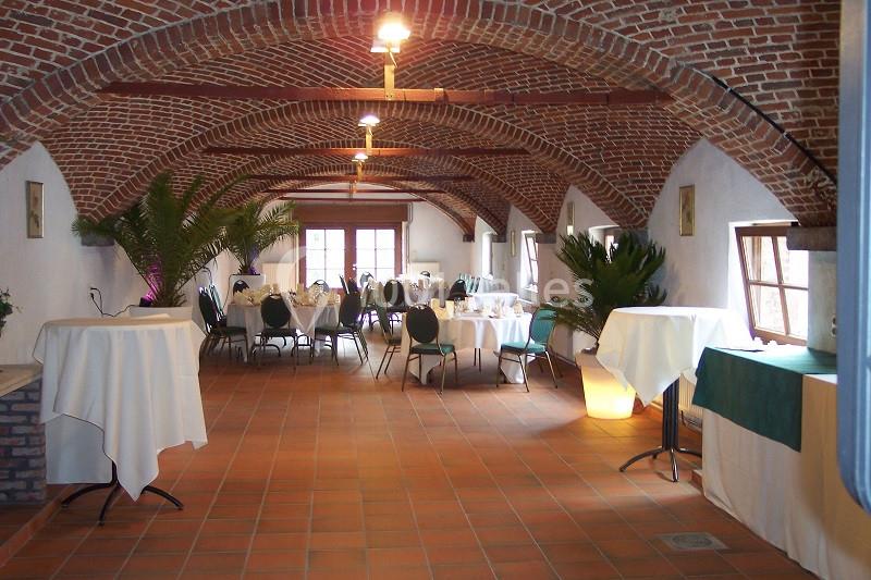 Salle voûtée en briques avec tables dressées et plantes décoratives, éclairée par des fenêtres et des lampes suspendues.