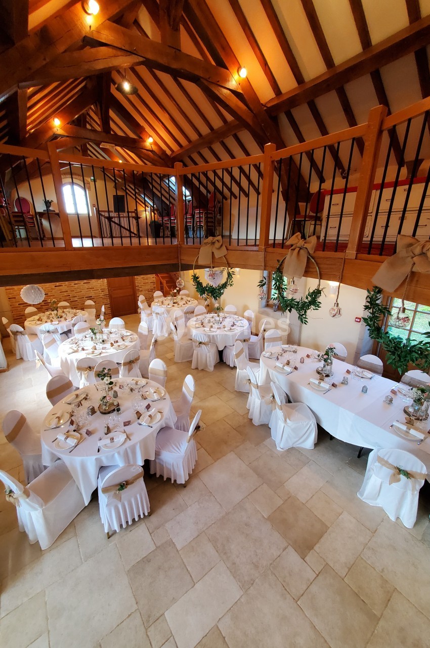 Salle de réception décorée avec des tables rondes dressées, chaises blanches et poutres apparentes en bois.