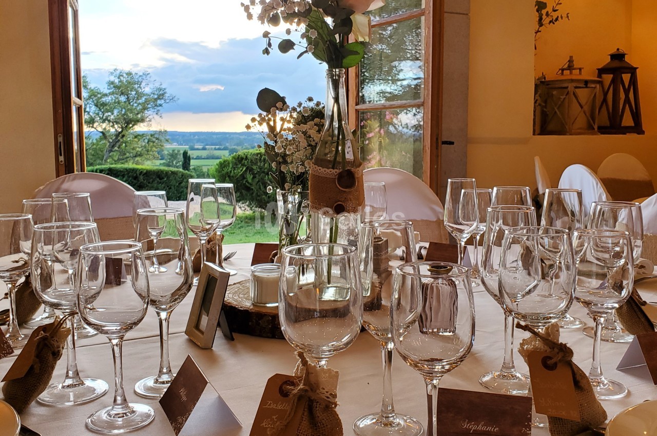 Table élégamment dressée avec des verres et décorations florales, vue sur un paysage verdoyant par une fenêtre ouverte.