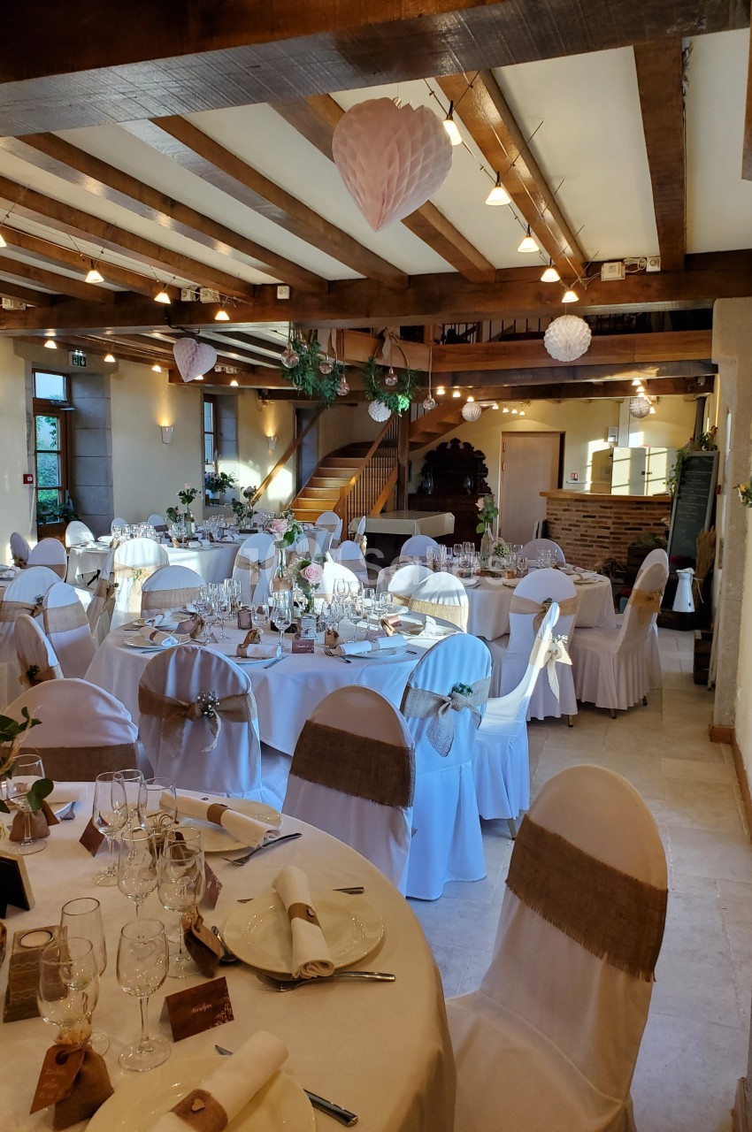 Salle de réception décorée avec des tables rondes dressées, chaises blanches et poutres apparentes au plafond.