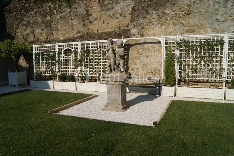 Statue en pierre sur un socle dans un jardin avec pelouse, gravier blanc et treillis végétalisé devant un mur en pierre.