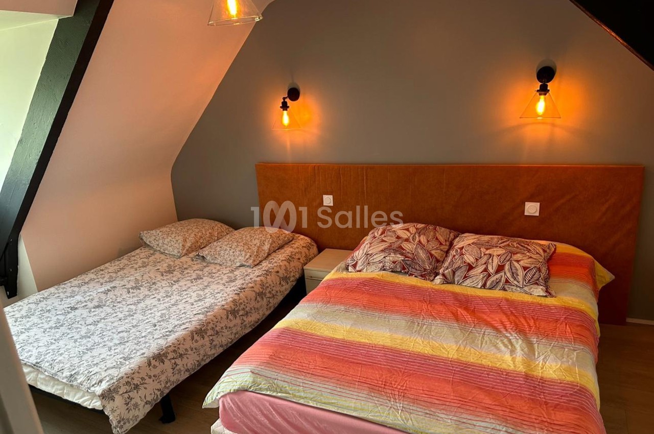 Deux lits dans une chambre mansardée avec mur gris, éclairage mural et parquet clair.