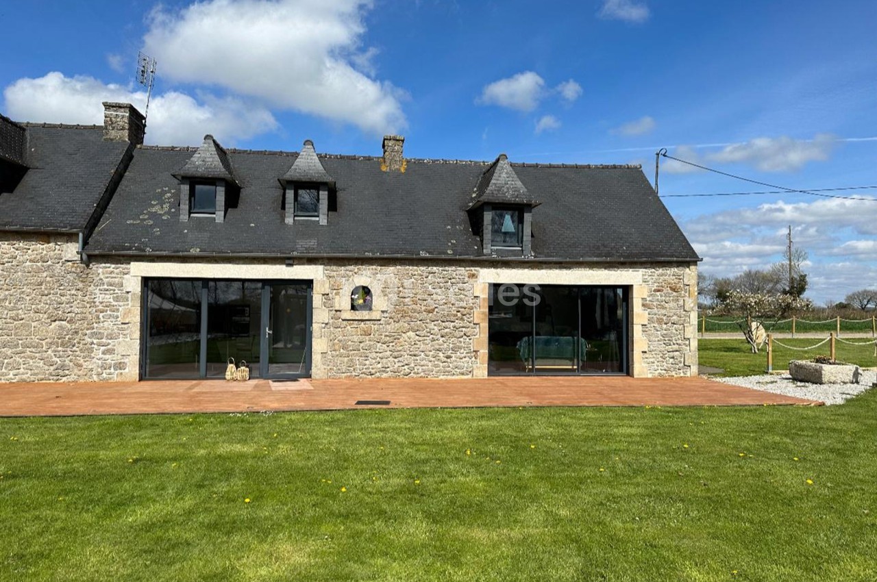 Façade d'une maison en pierre avec toit en ardoise, grandes baies vitrées et pelouse au premier plan.