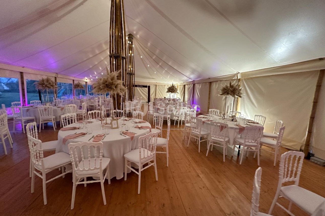 Salle de réception sous une tente avec tables rondes dressées, chaises blanches et éclairage tamisé.