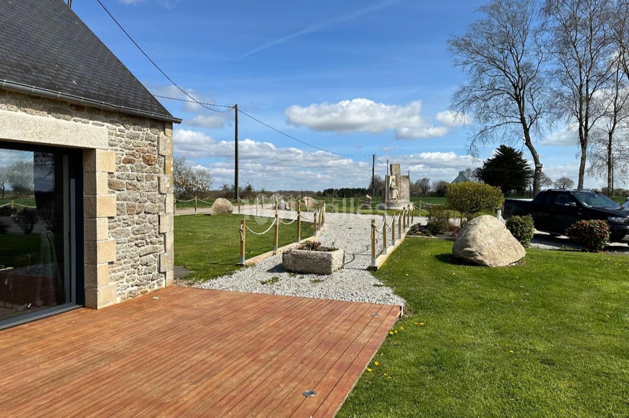 Allée en gravier bordée de poteaux en bois, menant à un jardin avec pelouse, arbres et rochers sous un ciel dégagé.
