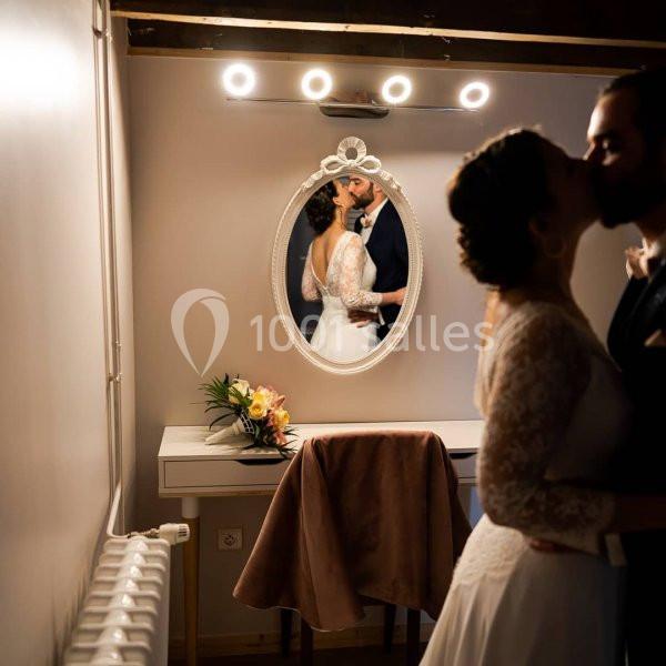 Un couple s'embrasse devant un miroir éclairé, reflétant la mariée en robe blanche et un bouquet posé sur une coiffeuse.