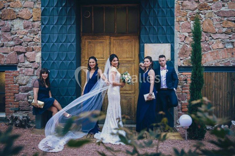 Une mariée en robe blanche pose avec quatre invités élégamment vêtus devant une porte en bois et un mur en pierre.