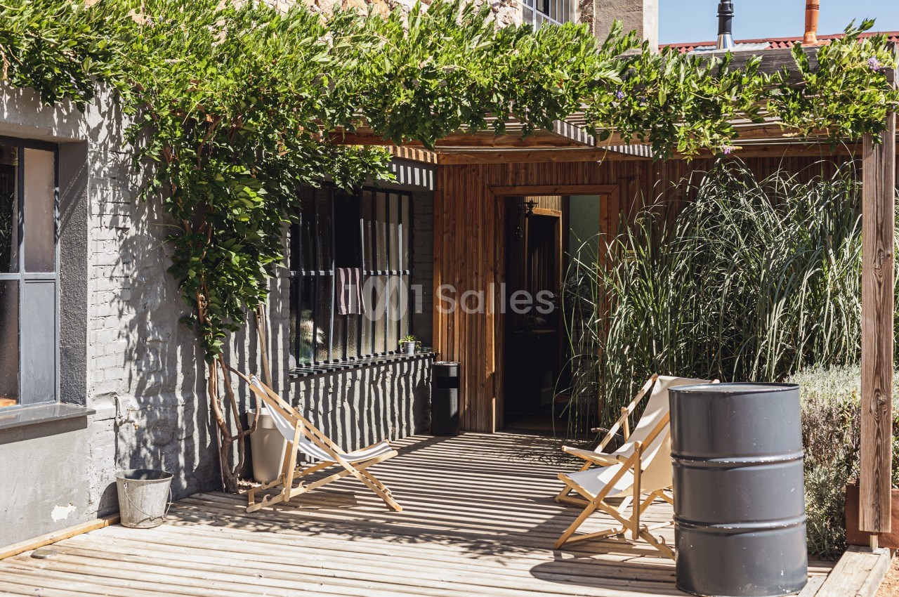 Terrasse en bois avec chaises longues, pergola couverte de verdure et plantes entourant l'entrée d'une maison.