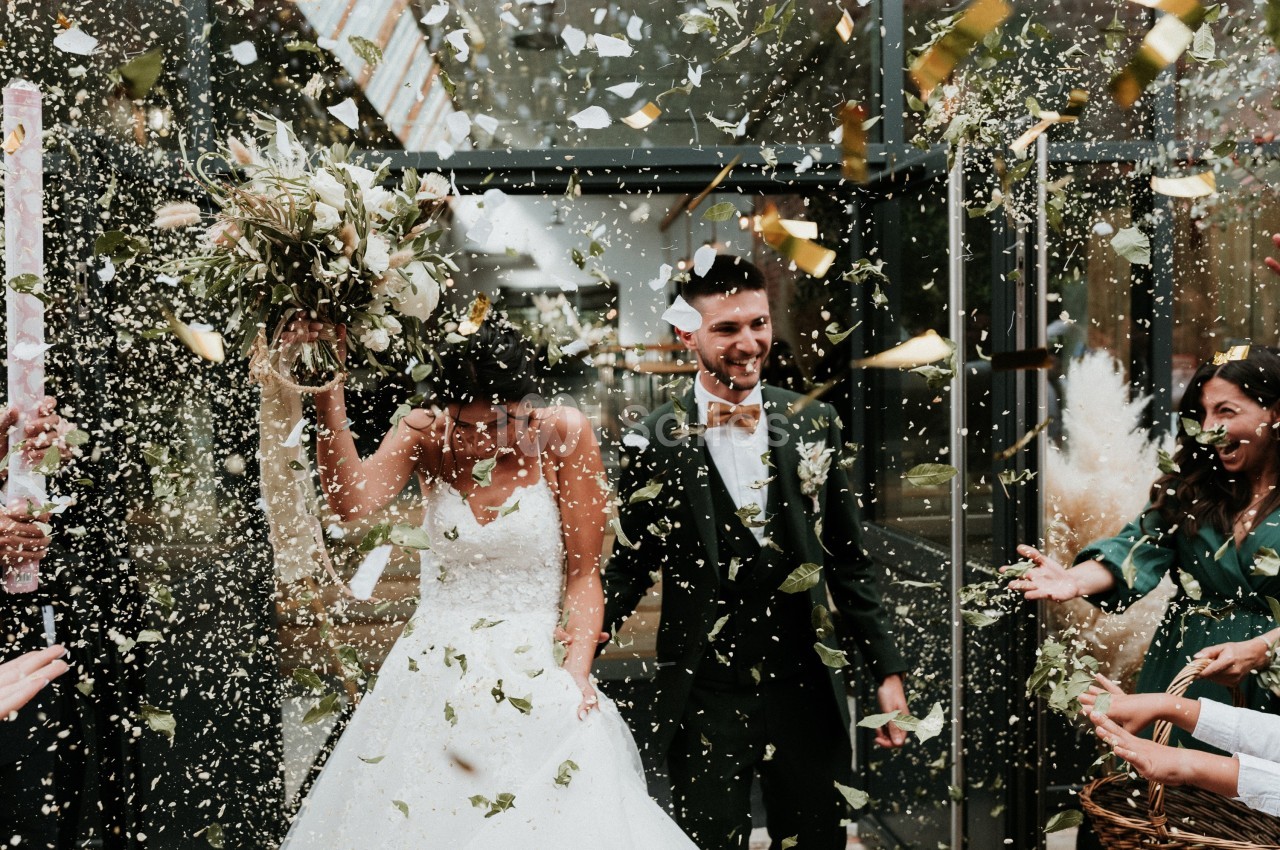 Un couple de mariés souriants sort sous une pluie de confettis, entouré d'invités célébrant leur union.