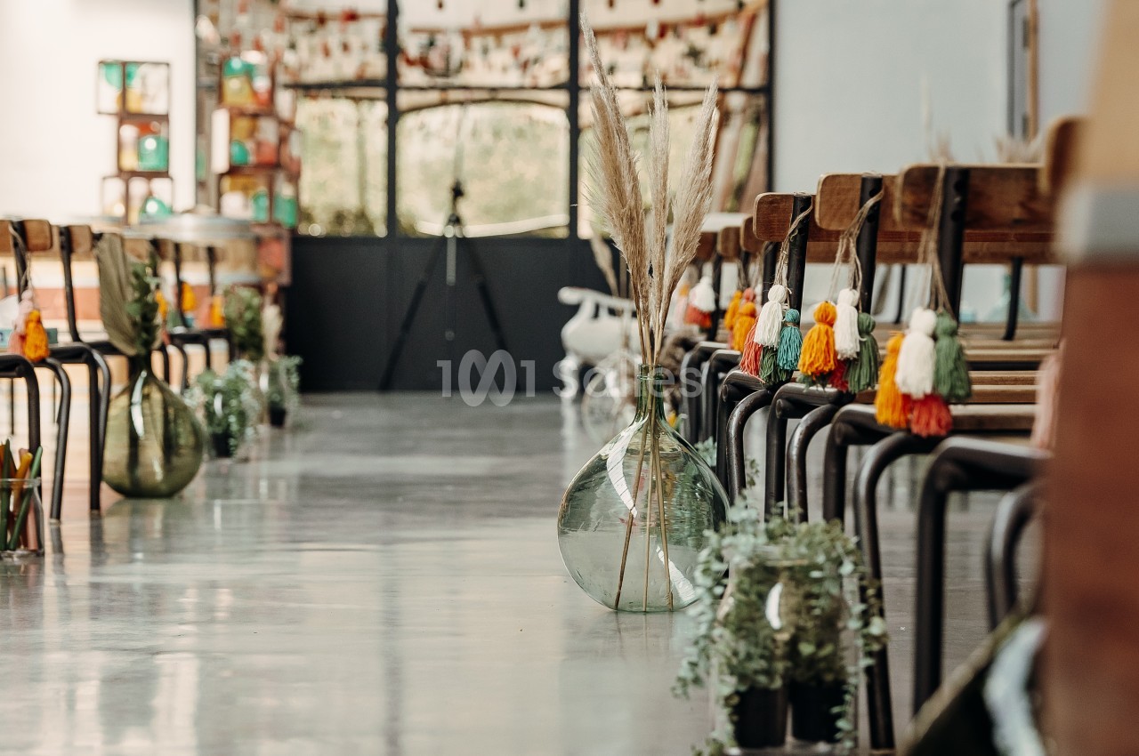 Allée décorée avec des vases en verre, des pampas et des guirlandes colorées, dans une salle lumineuse.