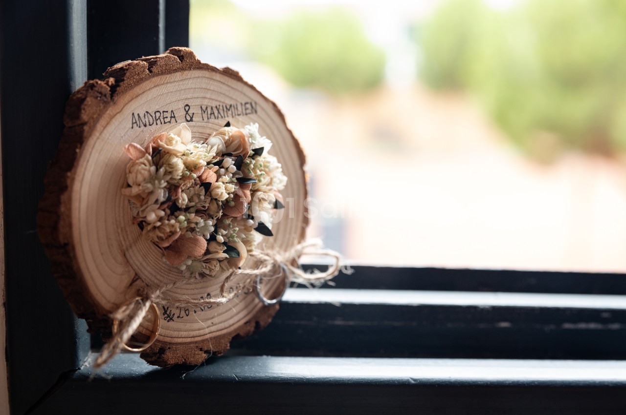 Tranche de bois gravée avec des prénoms, décorée de fleurs séchées, posée sur un rebord de fenêtre.