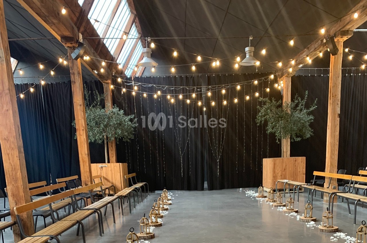 Salle décorée pour un événement avec guirlandes lumineuses suspendues, bancs en bois et lanternes alignées au sol.