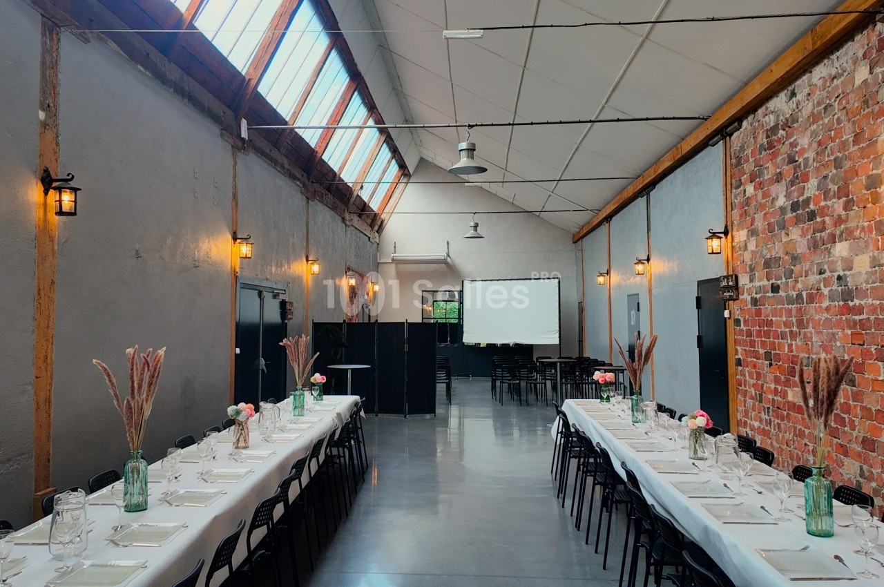 Salle de réception lumineuse avec tables dressées, décor rustique, murs en briques et éclairage suspendu.