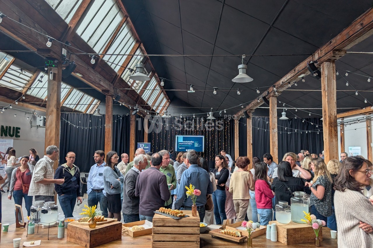 Groupe de personnes échangeant dans une grande salle avec toit vitré, tables en bois et buffet au premier plan.