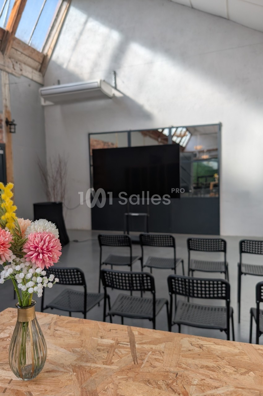 Salle lumineuse avec des chaises alignées face à un écran, vue depuis une table avec un vase de fleurs colorées.