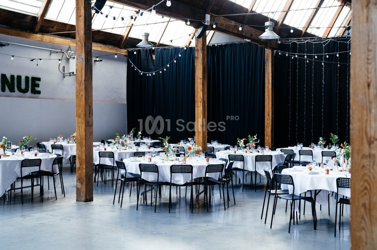 Salle de réception lumineuse avec tables rondes dressées, chaises noires et décorations florales.