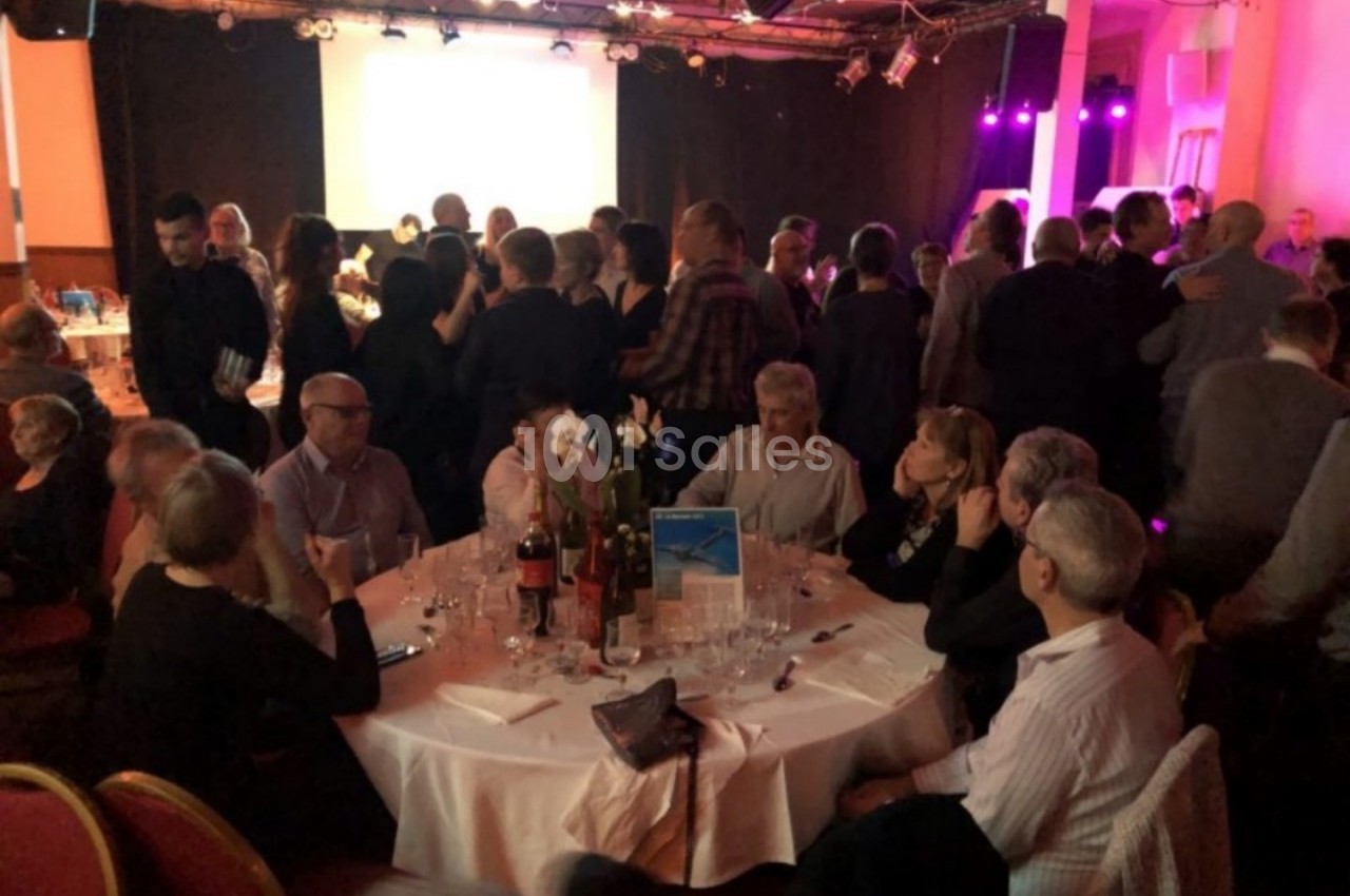 Des convives assis et debout dans une salle éclairée, autour de tables dressées pour un événement festif.