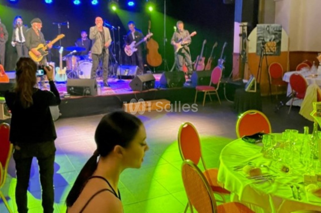 Groupe de musiciens jouant sur scène dans une salle éclairée, avec des tables dressées au premier plan.