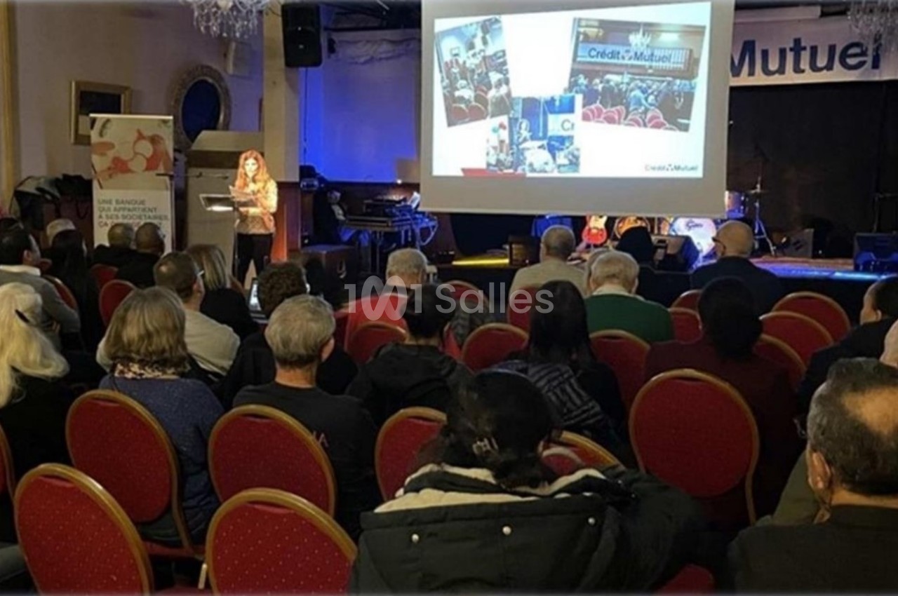 Une conférencière présente un diaporama à un public assis dans une salle avec des chaises rouges et une scène en arrière…