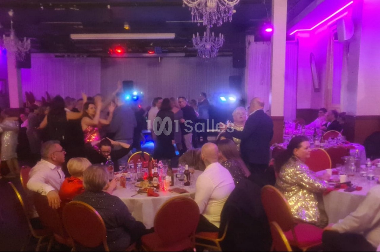 Salle de réception animée avec des invités assis à des tables et d'autres dansant sous des lumières colorées.