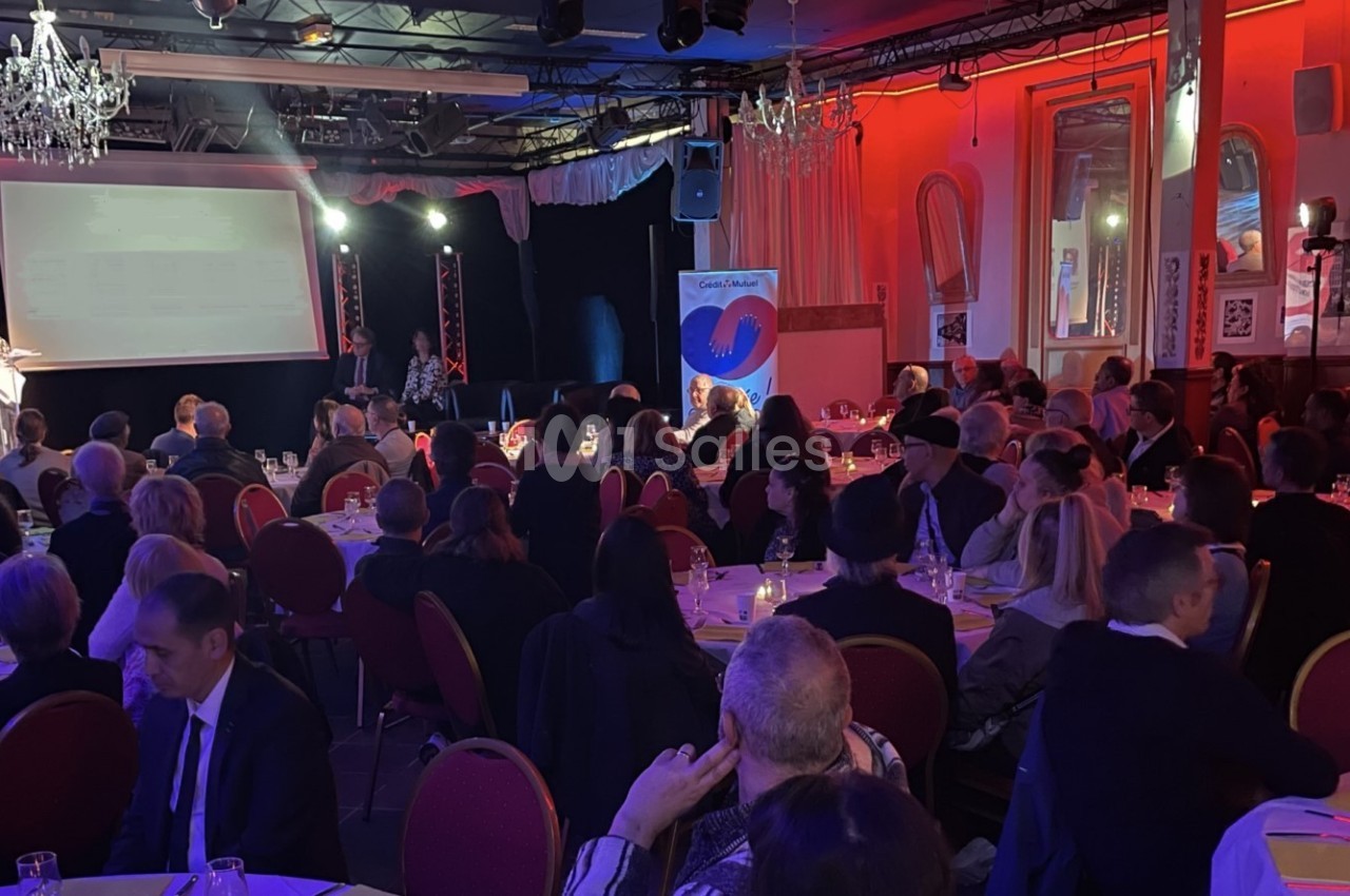 Salle de réception avec des invités assis à des tables rondes, éclairage tamisé et présentation sur scène.
