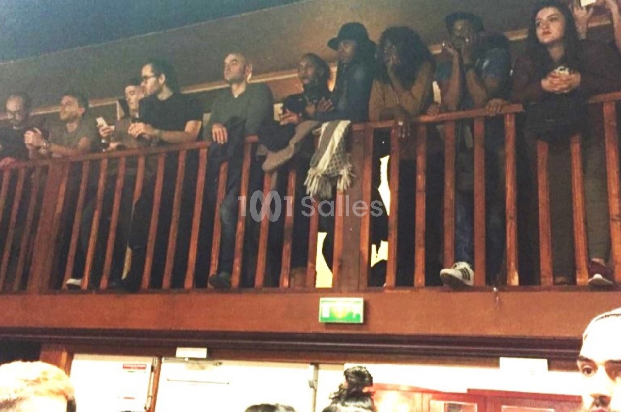 Des personnes debout sur une mezzanine en bois, observant une scène en contrebas dans un espace intérieur.