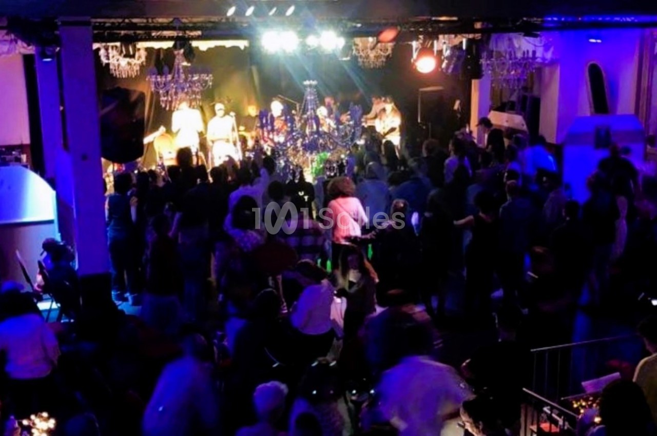 Salle sombre avec un public debout face à une scène éclairée où des musiciens jouent sous des projecteurs colorés.