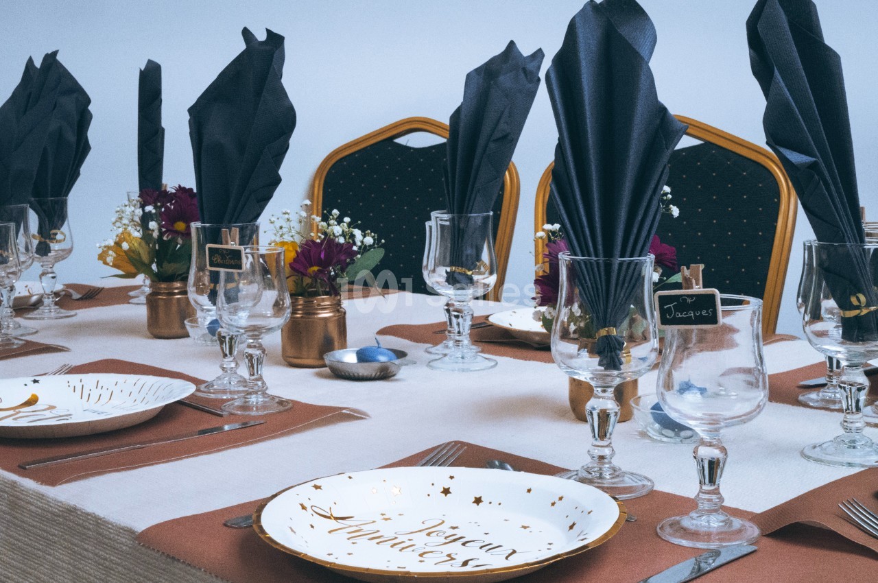 Table dressée avec vaisselle élégante, serviettes noires pliées, fleurs décoratives et marque-places sur fond clair.