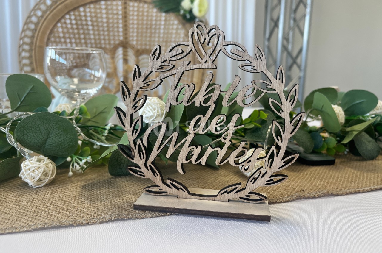 Panneau en bois gravé ’Table des Mariés’ entouré d'une couronne végétale, posé sur une table décorée.