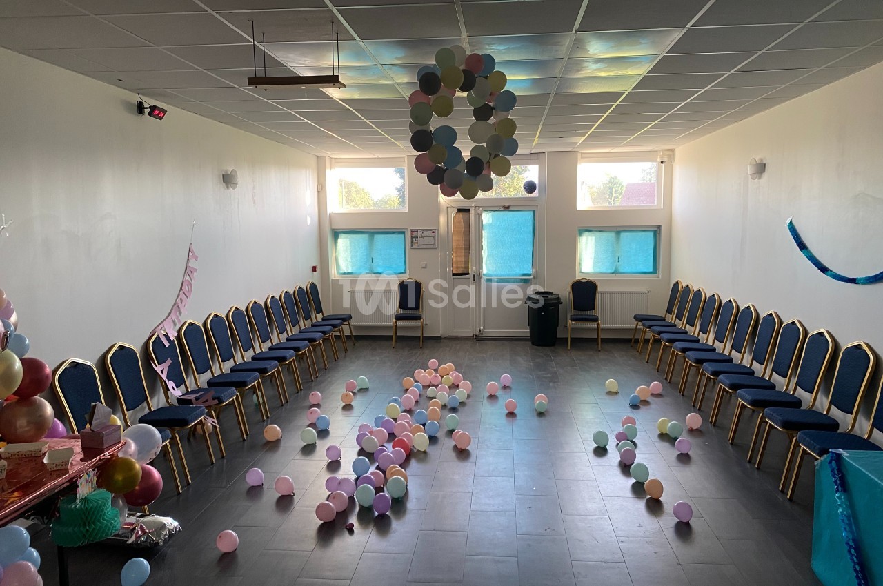 Salle décorée pour une fête avec des ballons au sol, des chaises alignées le long des murs et des guirlandes suspendues.