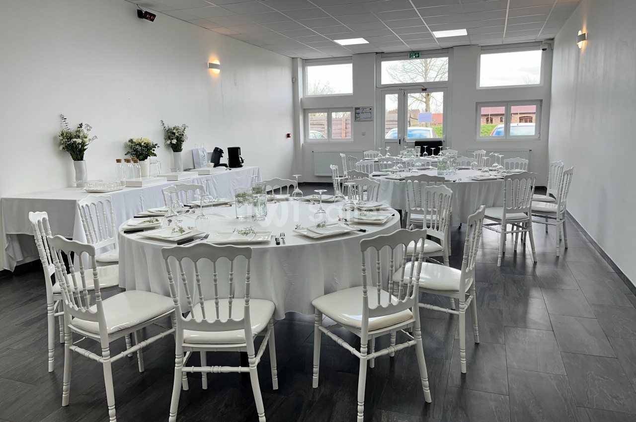 Salle de réception lumineuse avec tables rondes dressées, chaises blanches et décorations florales.