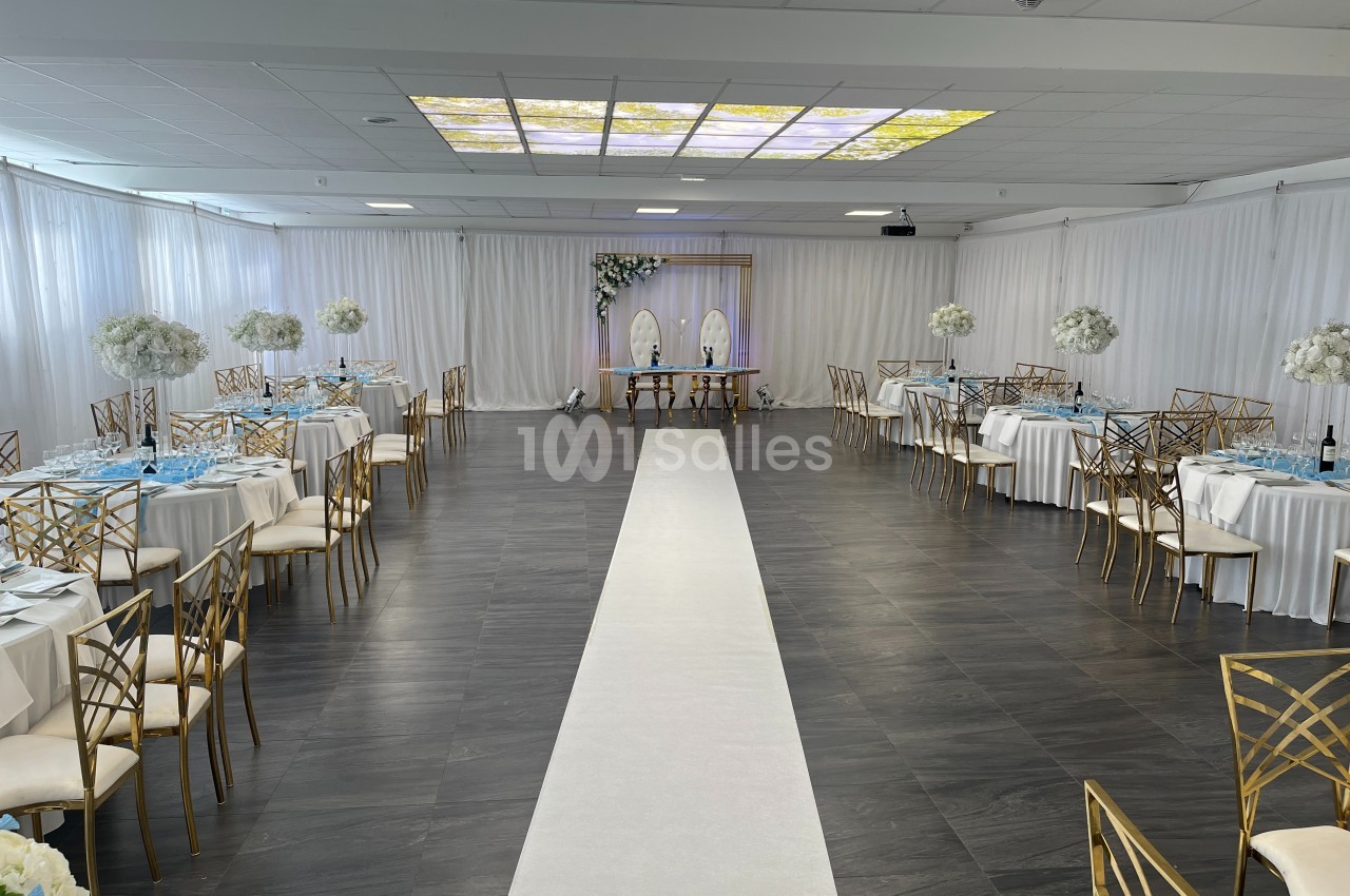 Salle de réception décorée avec des tables rondes, des chaises dorées, un tapis blanc central et un autel fleuri au fond.
