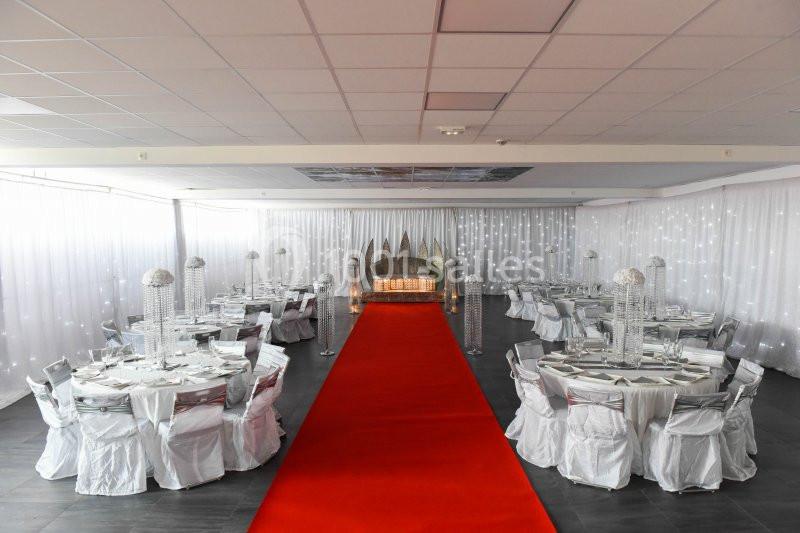 Salle de réception décorée avec des tables rondes, chaises blanches et un tapis rouge menant à une estrade ornée.