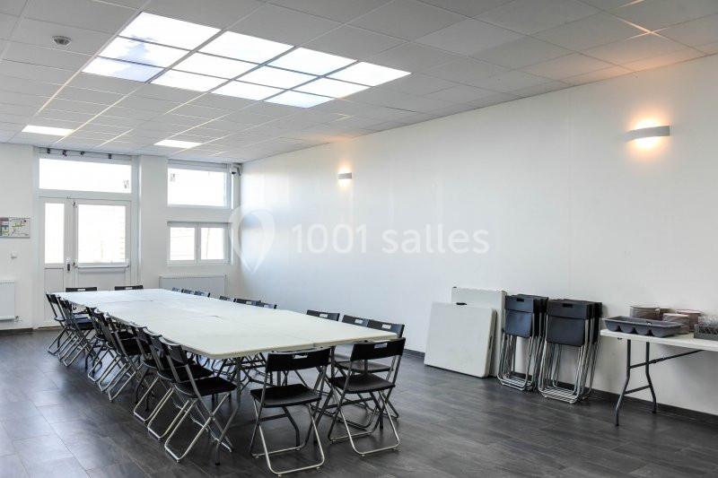 Salle lumineuse avec grandes fenêtres, équipée d'une longue table entourée de chaises pliantes et d'un coin rangement.