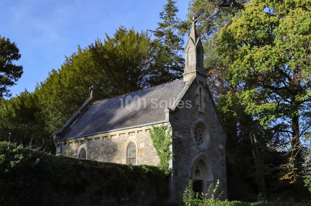 Petite chapelle en pierre entourée d'arbres, avec un toit en ardoise et une façade ornée d'une rosace.
