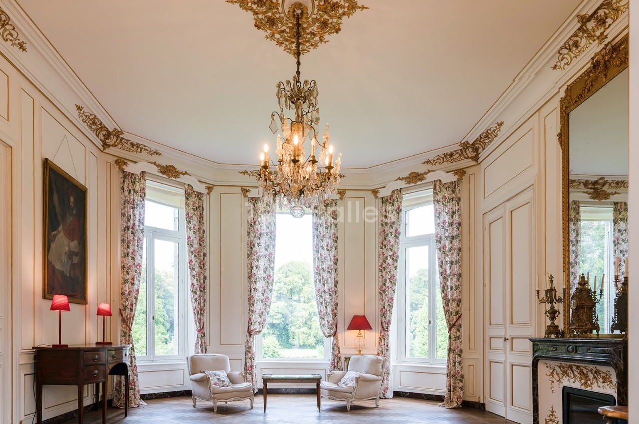 Salon élégant avec moulures dorées, lustre central, grandes fenêtres, rideaux fleuris et mobilier classique.