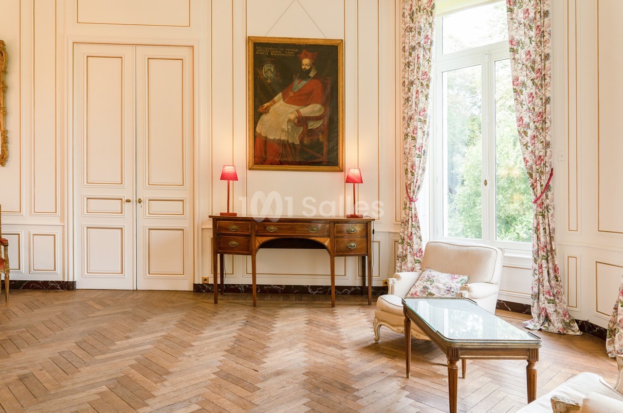 Salon élégant avec parquet en bois, mobilier classique, tableau ancien, grandes fenêtres et rideaux fleuris.