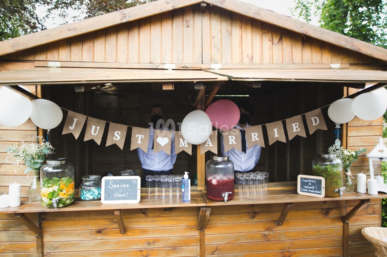 Stand en bois décoré pour un mariage, avec une guirlande ’Just Married’, boissons et bonbonnières disposées devant.